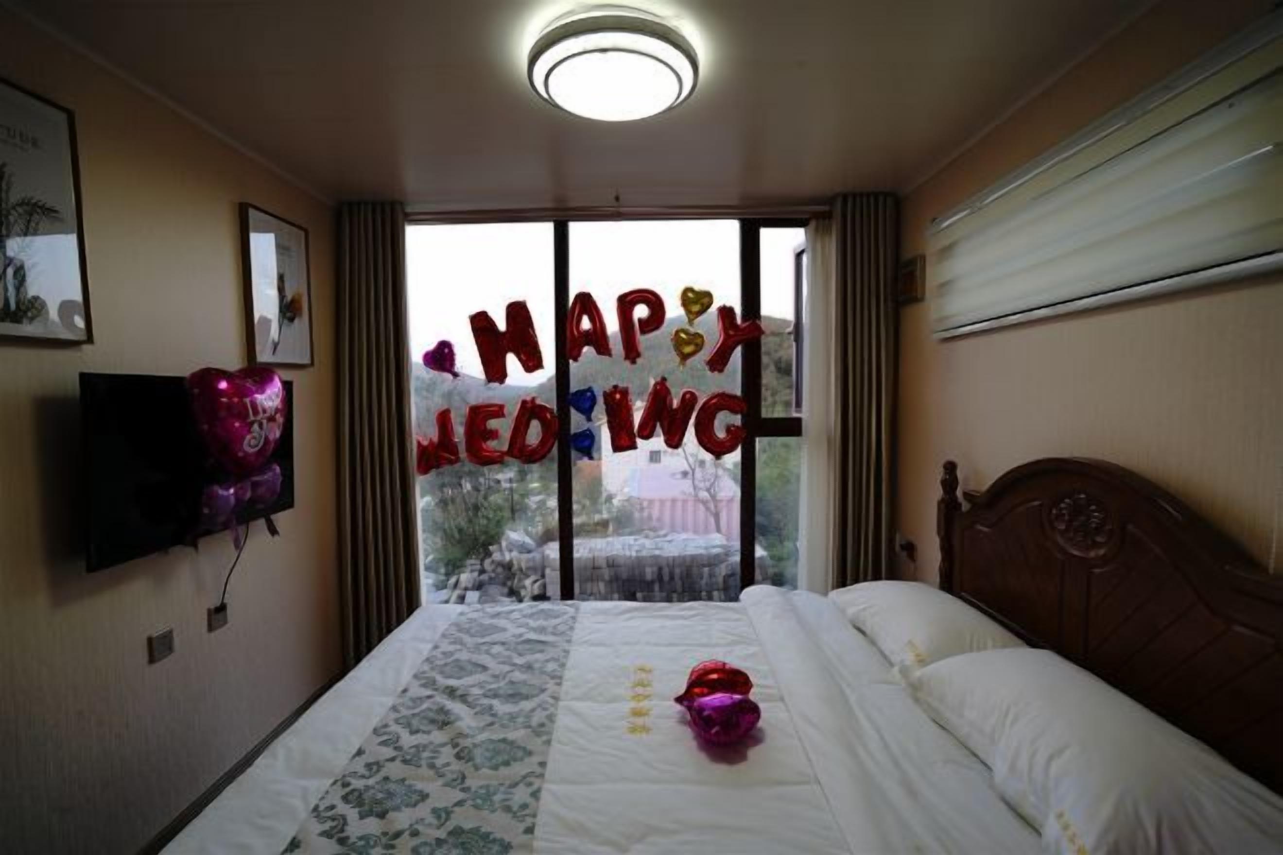 Una habitación de hotel con una cama, un televisor de pantalla plana, un ventanal con vistas y una decoración que dice 'HAPPY WEDDING'.