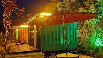 An outdoor patio area with a wooden deck, metal chairs, a round table, and a large umbrella.