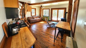 A spacious kitchen with wooden flooring, a dining area, a sofa, and a large window offering a view of the outdoors. - Cabin Retreat in Southern Vermont (Putney)