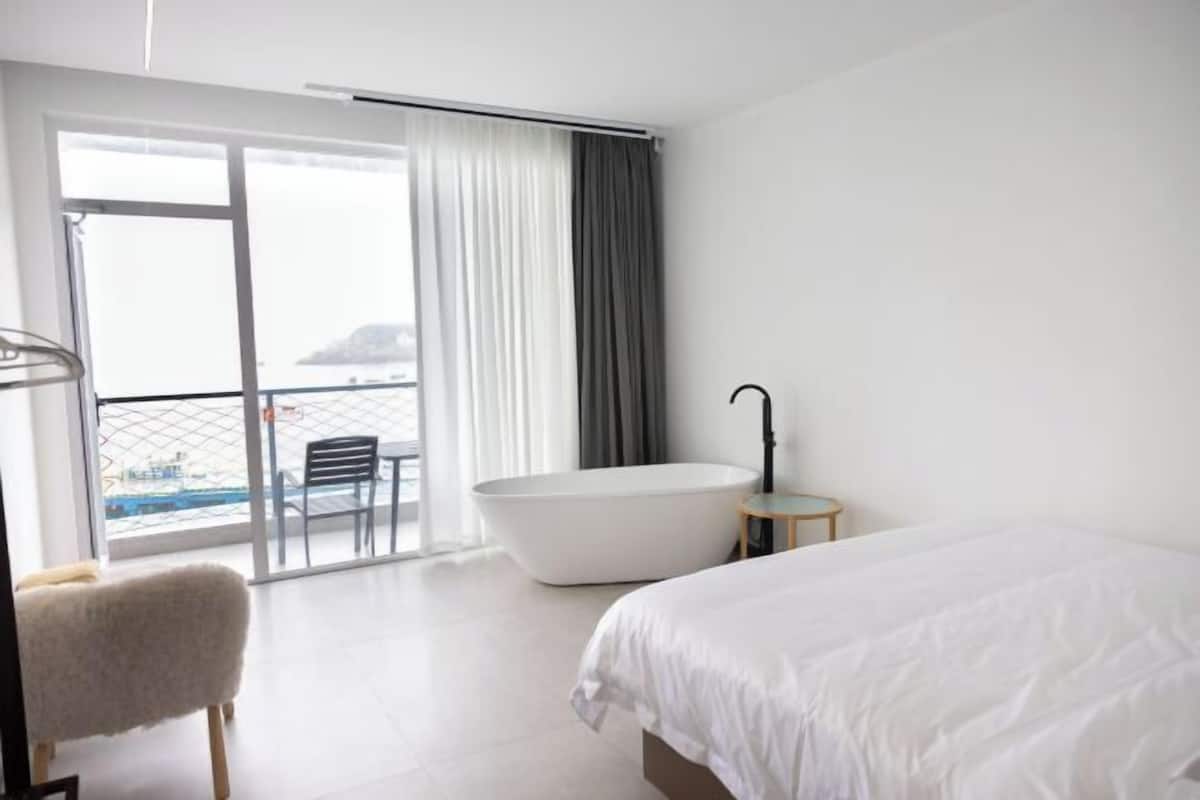 Ein modernes Hotelzimmer mit einem großen Bett, einer Badewanne, einem Sessel und Meerblick durch eine Schiebetür.