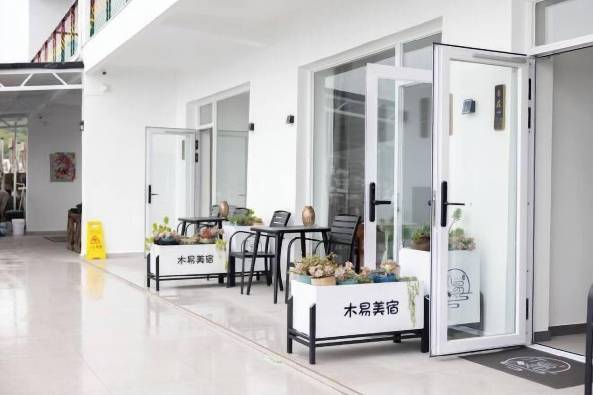 Eine moderne Hotelhalle mit klarem, minimalistischem Design. Es befinden sich zwei Rezeptionen mit Glastüren darin, jeweils mit einem kleinen Blumentopf auf dem Tisch. Der Boden ist mit Fliesen ausgelegt, und links ist ein gelbes Warnschild sichtbar.