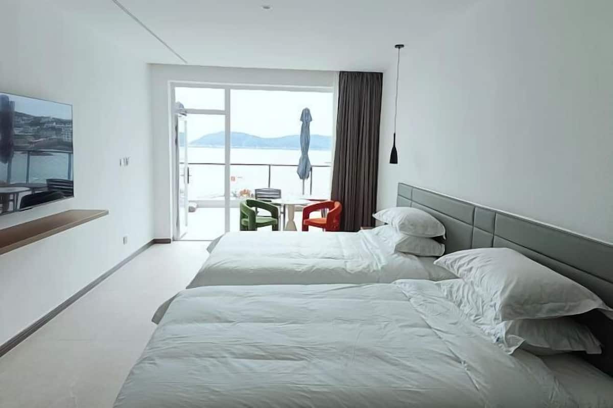 Ein Hotelzimmer mit zwei Betten, einem an der Wand befestigten Fernseher, einem Balkon mit Meerblick und einem kleinen Tisch mit Stühlen.