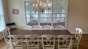 Een eetkamer met een grote houten tafel en witte stoelen, een kroonluchter en uitzicht door een raam met jaloezieën.