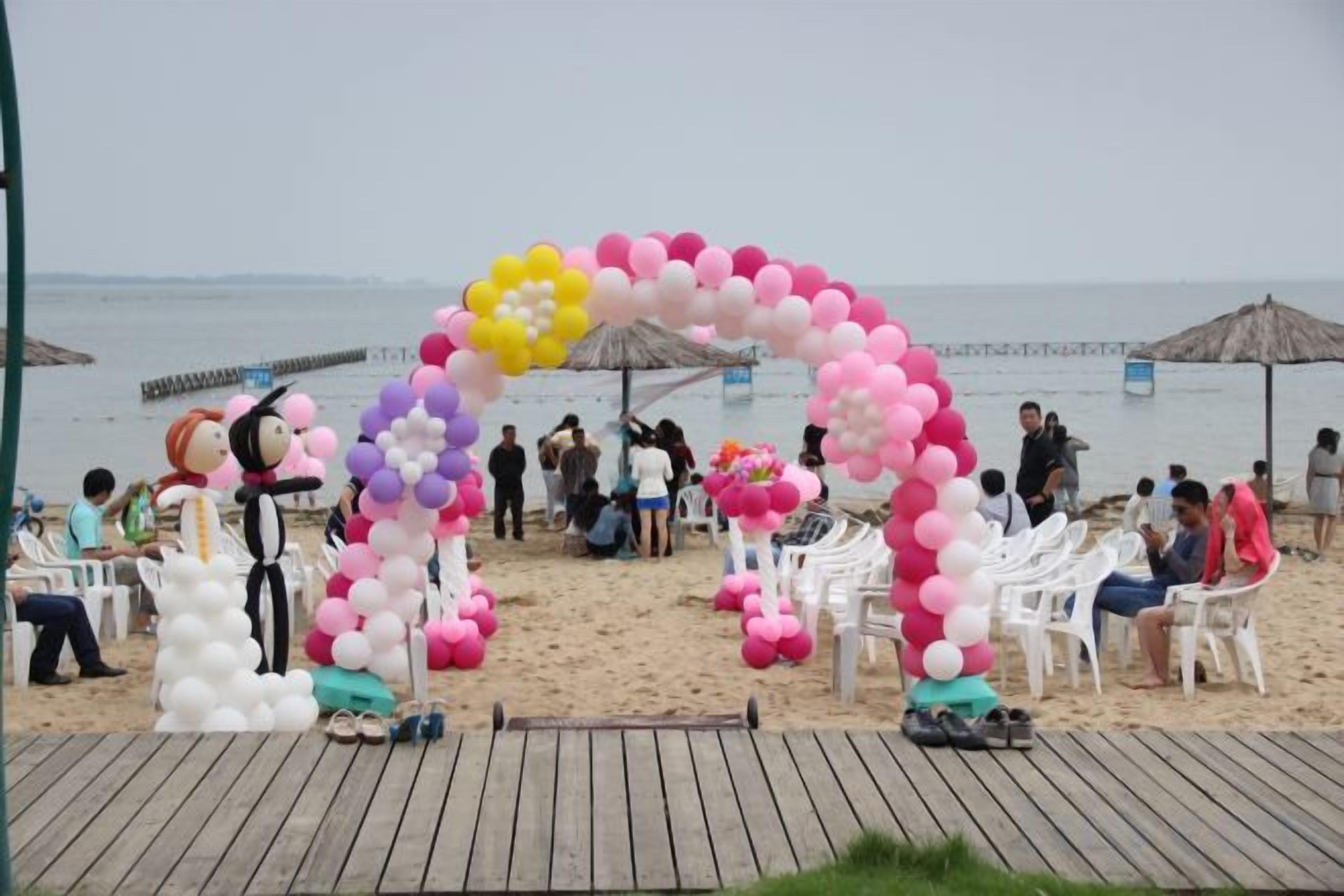 Escena de playa con terraza de madera, sillas blancas y un arco de globos.