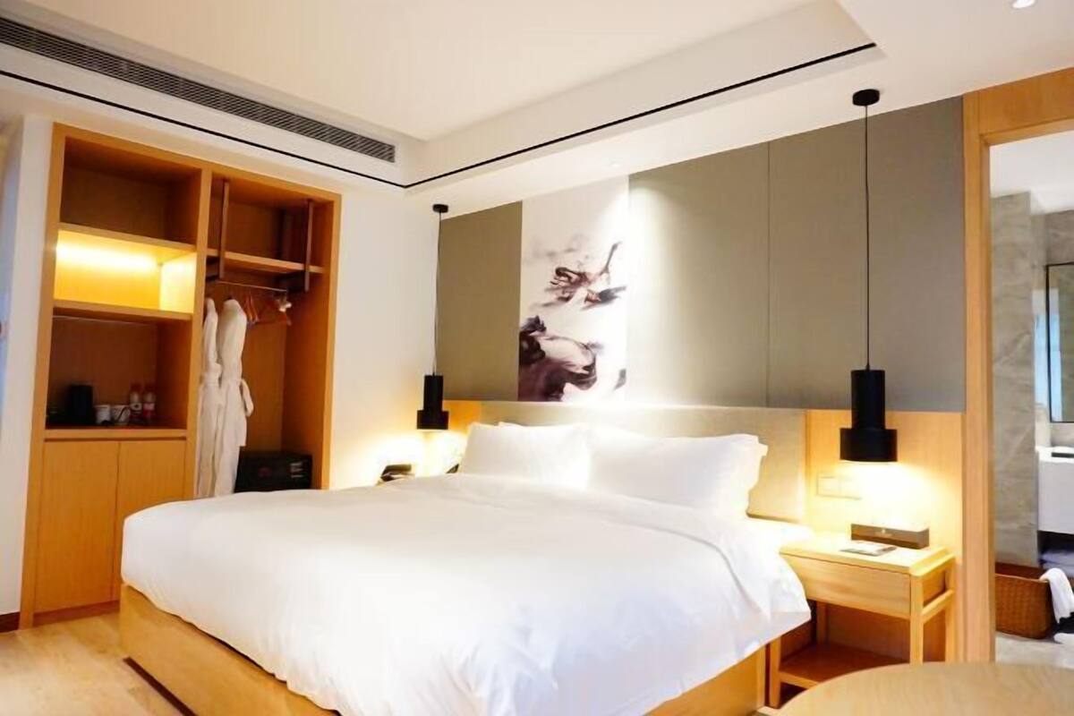 Una habitación de hotel moderna con una cama grande, un armario, una mesita de noche con una lámpara y una obra de arte colgada en la pared.