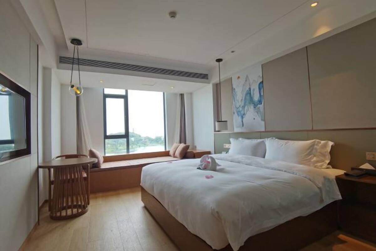 Una habitación de hotel moderna con una cama grande, una mesita pequeña, un rincón con asiento junto a la ventana y un cuadro en la pared.
