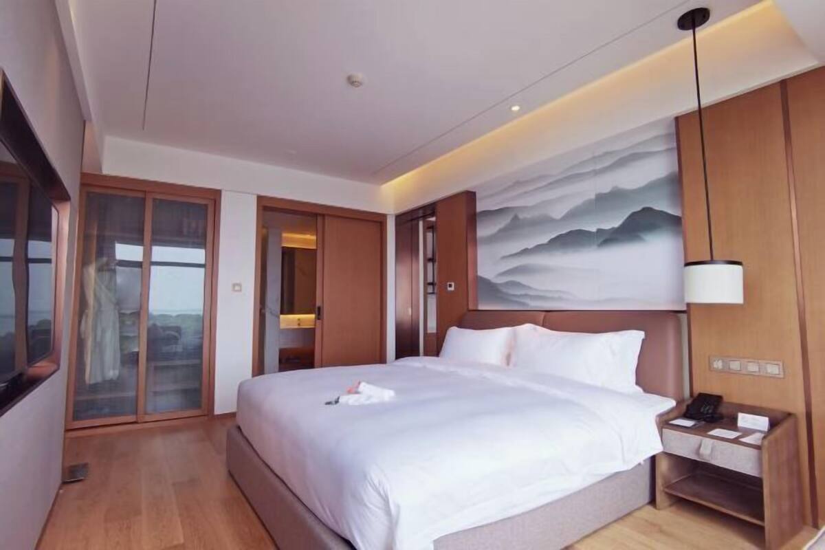 Una habitación de hotel moderna con una cama grande, una mesita de noche, una lámpara de pared y una puerta corredera que da a un balcón.