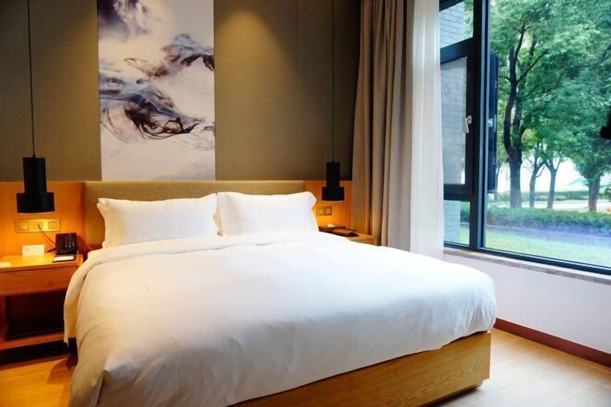 Una habitación de hotel moderna con una cama grande, cabecera de madera y vistas a árboles a través de un ventanal.