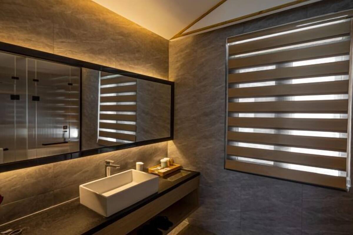 Un baño moderno con un espejo grande, un lavamanos blanco y una ventana con persianas horizontales.