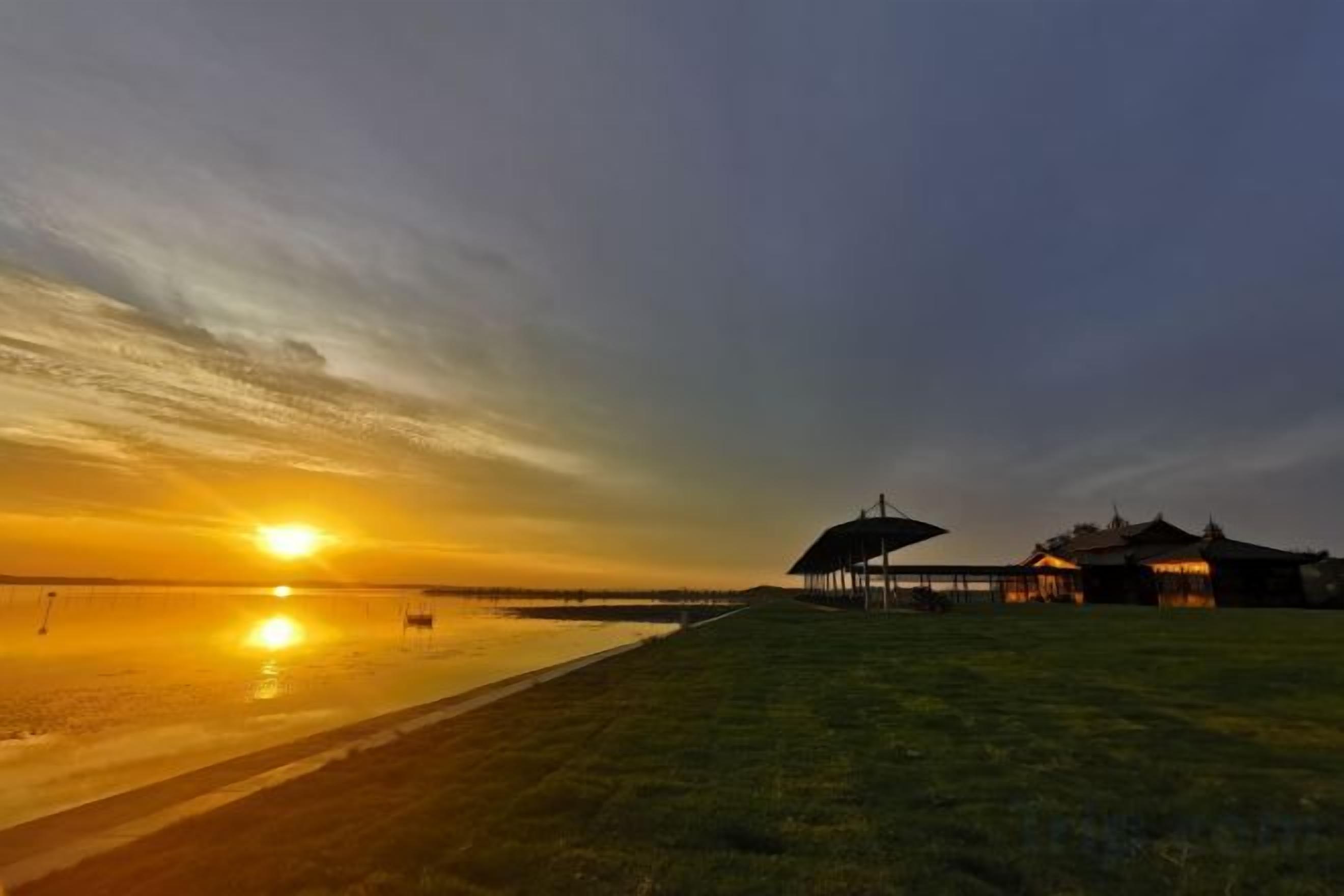 Un coucher de soleil sur une plage, avec une zone herbeuse et un bâtiment doté d’une structure couverte.