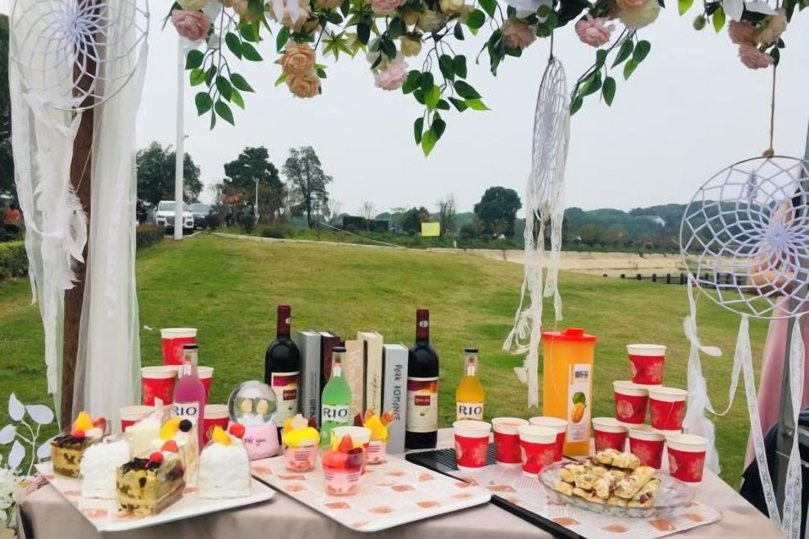 Mesa al aire libre con diferentes alimentos y bebidas, rodeada de elementos decorativos.
