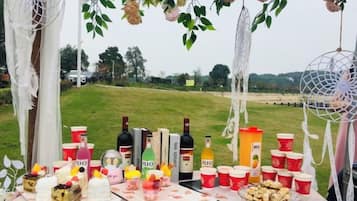 Mesa al aire libre con diferentes alimentos y bebidas, rodeada de elementos decorativos.