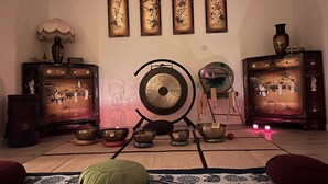 Een kamer met een meditatieruimte, inclusief een gong, diverse kommen en een mat. Er zijn wanddecoraties en traditionele kasten met decoratieve objecten.