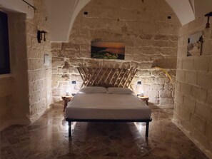 A stone-walled bedroom with a bed, two bedside tables with lamps, and a painting on the wall. - HUG HOUSE (Giuggianello)