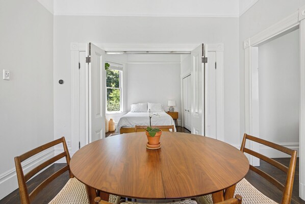 Une pièce comprenant une table ronde en bois, deux chaises en bois, un lit avec une literie blanche, une table de chevet avec une lampe et une fenêtre donnant sur un paysage verdoyant.