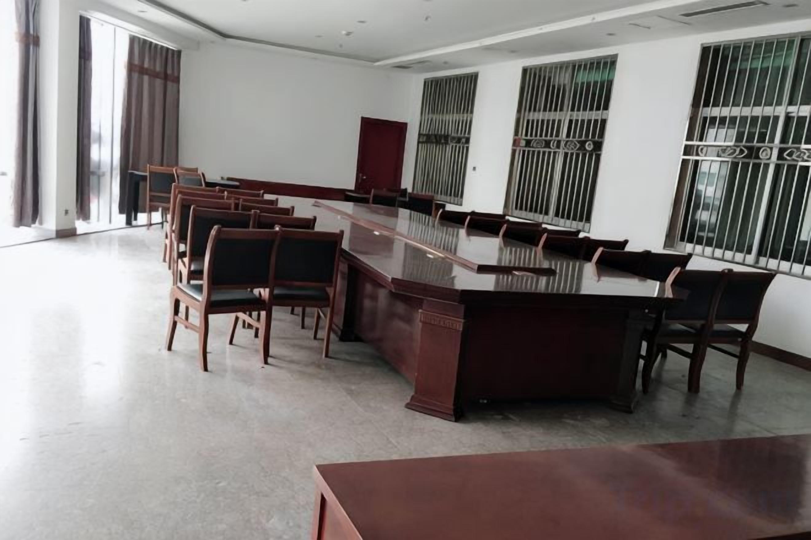 Un salón de conferencias con una larga mesa y sillas, ventanas amplias con barrotes metálicos y un techo alto.