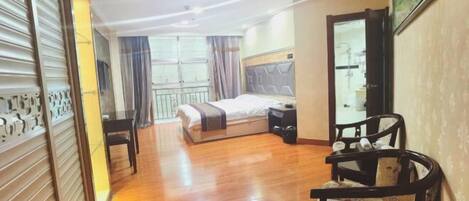A hotel room with a wooden floor, a bed with a headboard, a bedside table, a chair, and a large window with curtains.