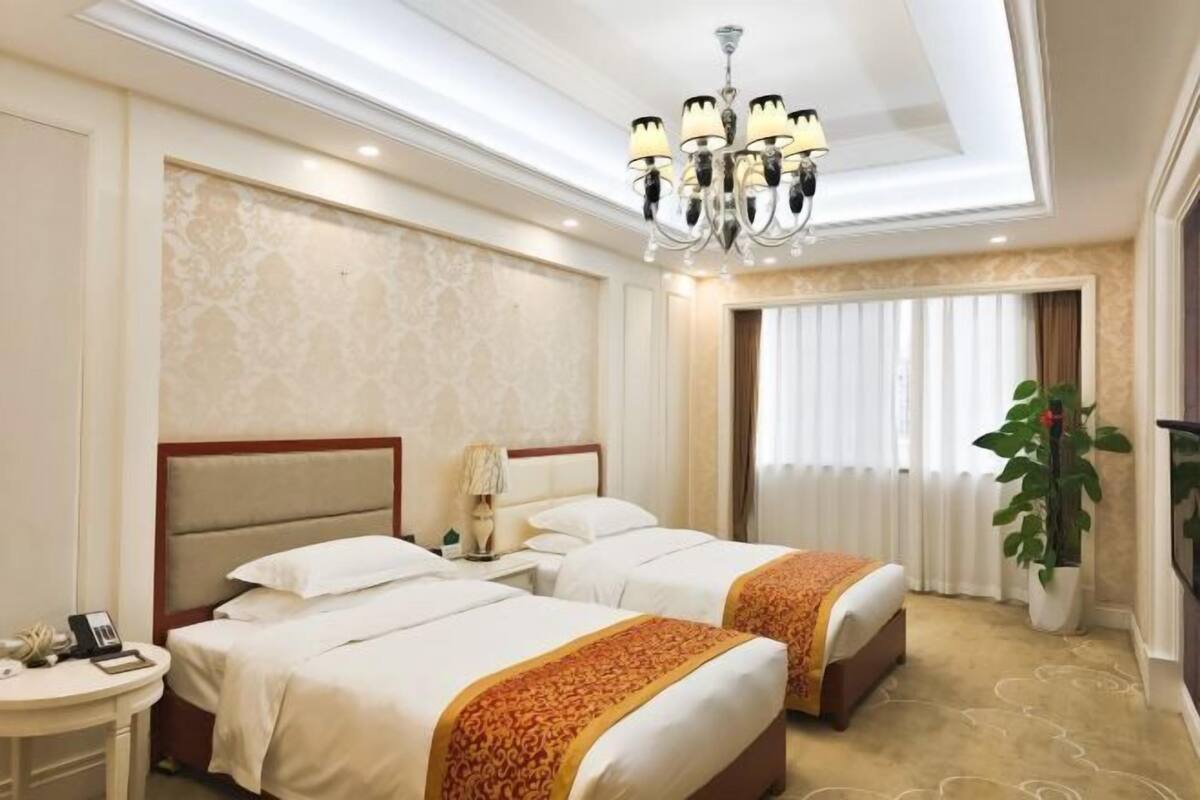 A hotel room with two beds, a chandelier, a plant, and a window with curtains.