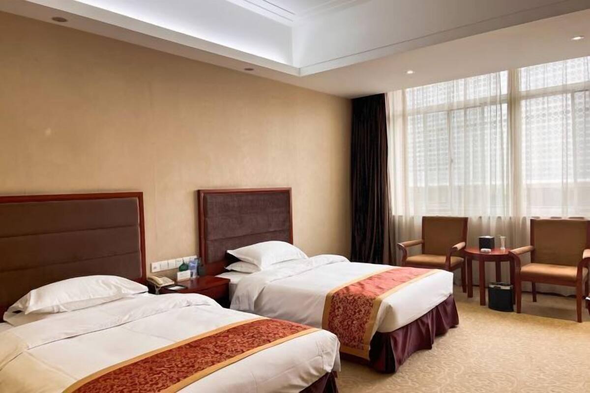 A hotel room with two beds, a desk, chairs, and a large window with curtains.