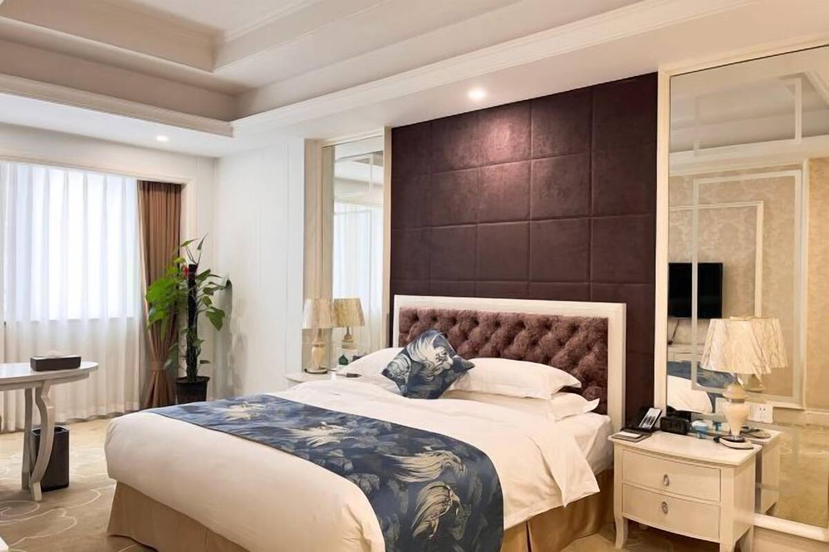 A hotel room with a large bed, a bedside table with a lamp, a mirror, and a window with curtains.
