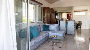 A living room with a glass coffee table, a sofa with patterned cushions, and a view of the kitchen through an open door. - Villa Rose (Anavissos)