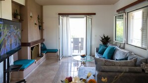 A living room with a TV, seating area, and a glass door leading to an outdoor space. - Villa Rose (Anavissos)