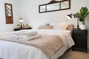 Una cama bien tendida con toallas dobladas, una lámpara de noche, una planta y un espejo en la pared.