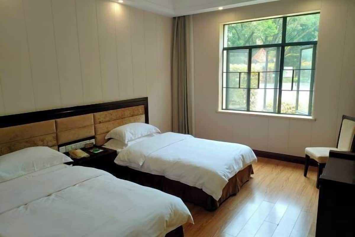 Quarto de hotel com duas camas, uma mesa de cabeceira, uma cadeira e uma janela grande com vista para o verde.
