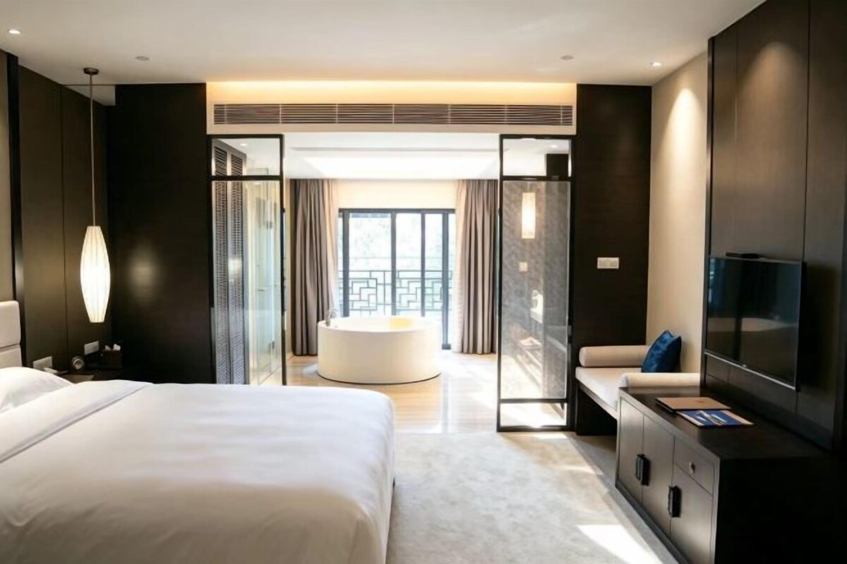 A modern hotel room with a large bed, a seating area with a sofa and a round table, a TV, and a balcony with curtains.