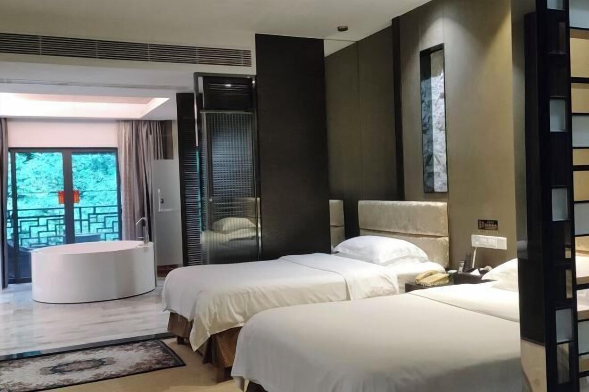 A hotel room with two beds, a round bathtub, a large window, and a view of greenery.