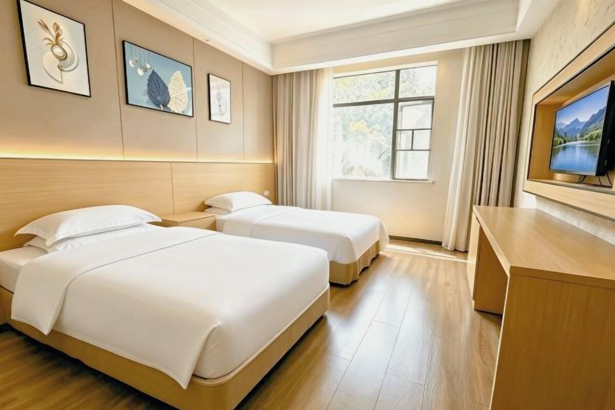A hotel room with two beds, a TV, a desk, and a window with curtains.