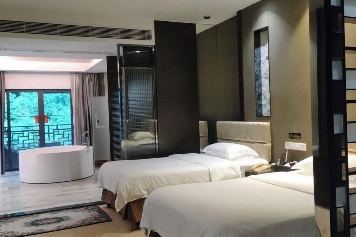 A hotel room with two beds, a round bathtub, and a large window with a view of greenery.