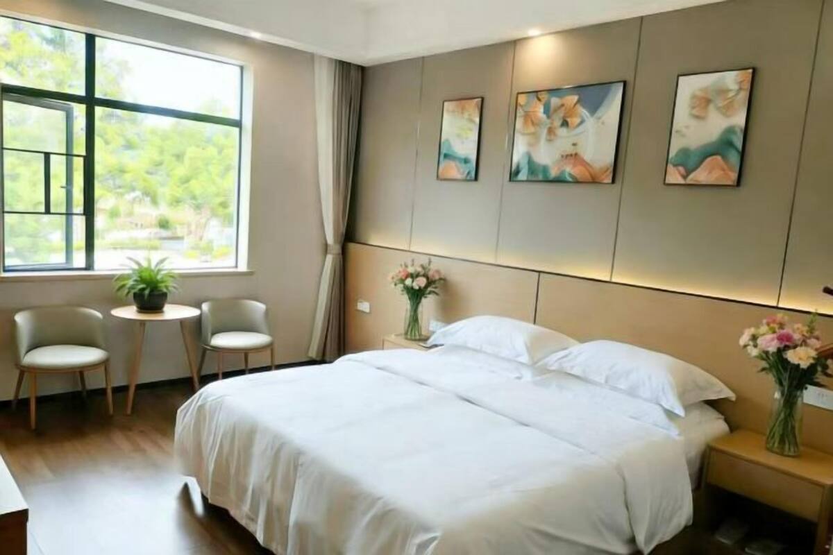 Quarto de hotel com uma cama grande, duas cadeiras, uma mesa pequena e uma janela com vista para o verde.