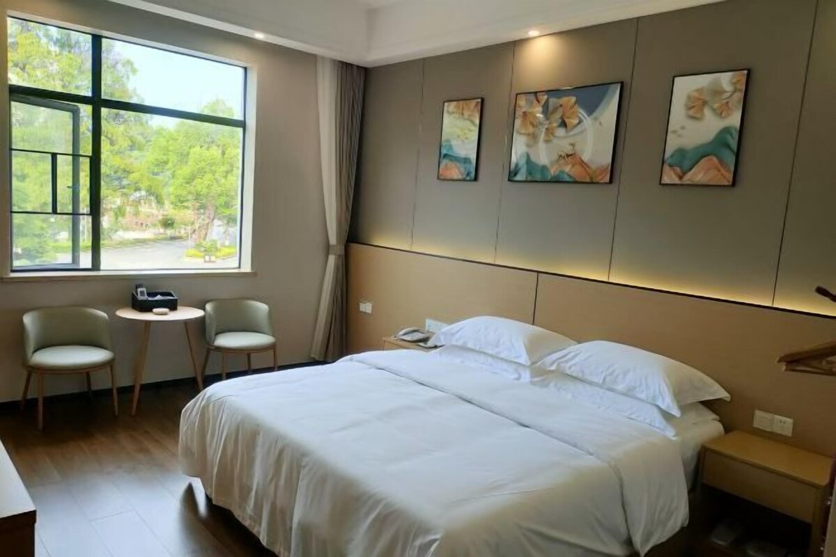 A hotel room with a large bed, two chairs, a small table, and a window with a view of greenery.