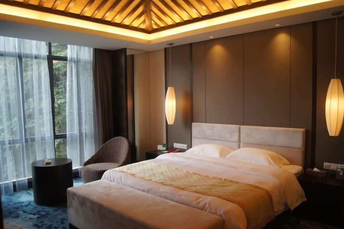 Quarto de hotel moderno com uma cama grande, uma cadeira, uma mesinha de cabeceira e vista para o verde pela janela.