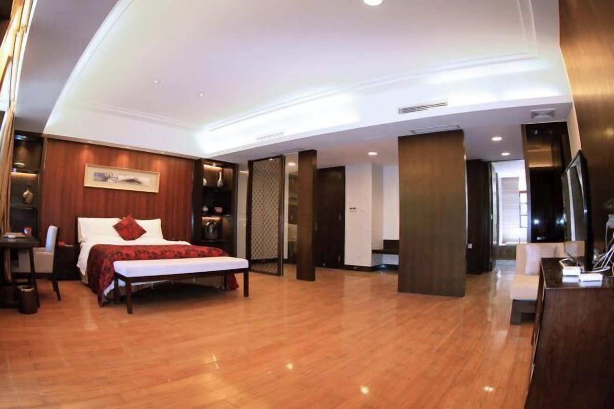 A modern hotel room with a wooden floor, a bed with a red accent pillow, a desk, and a television.