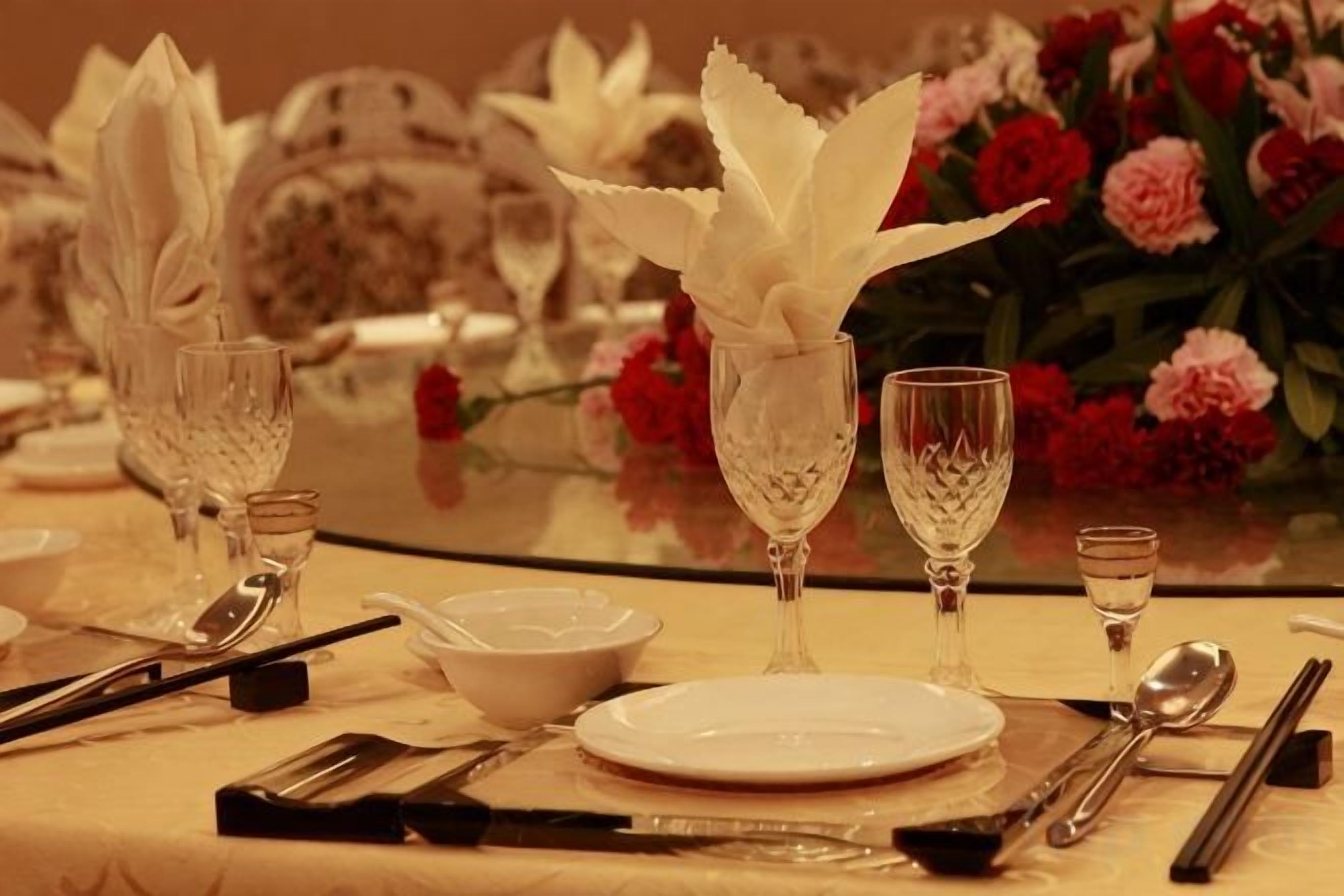Una mesa dispuesta con servilletas blancas, copas de vino y cubiertos, con un arreglo floral al fondo.