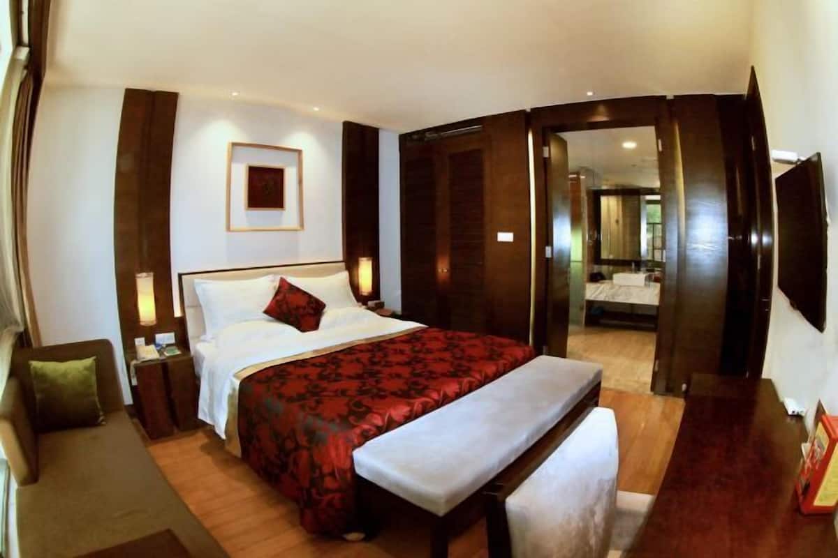 Quarto de hotel com uma cama grande, um banco, uma mesinha de cabeceira e uma televisão presa na parede.