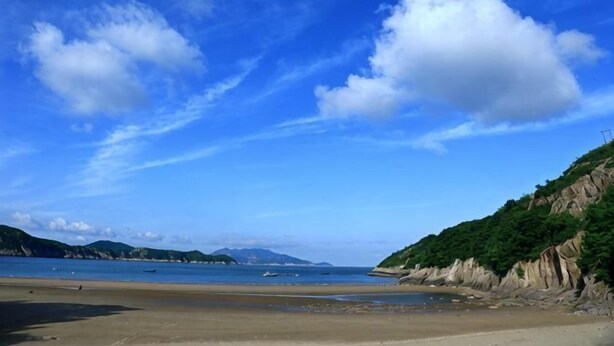 Eine Küstenlandschaft mit Strand, Klippen und fernen Bergen unter blauem Himmel.