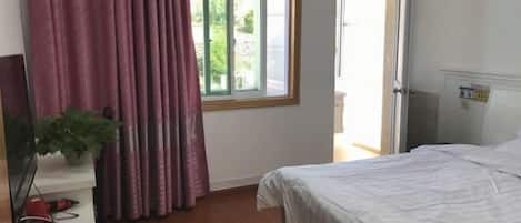 Quarto com cama, janela com cortinas, porta e uma pequena mesa com planta.