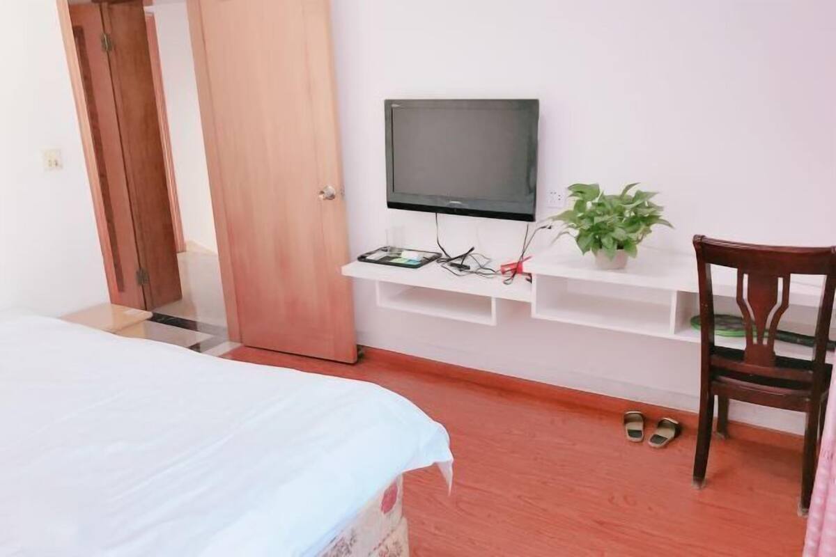 Une chambre à coucher avec un lit, un téléviseur fixé sur une étagère blanche, une porte en bois et une paire de chaussons posés sur le sol.