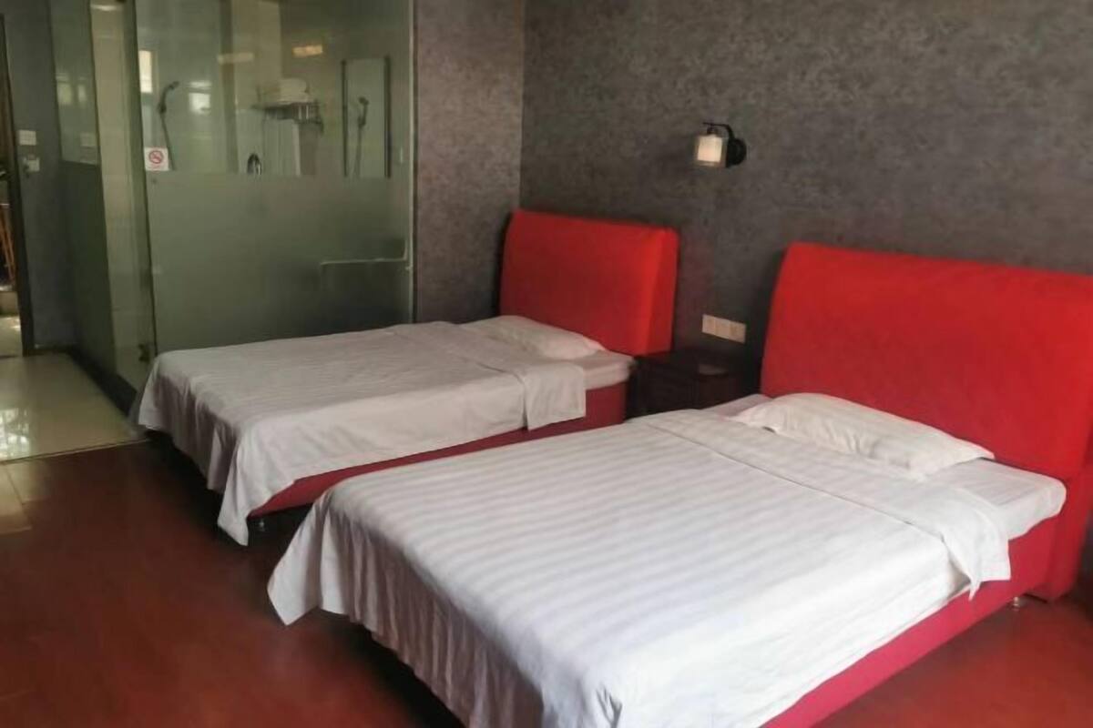 Une chambre d’hôtel avec deux lits, une tête de lit rouge, une porte en verre et un luminaire fixé au mur.