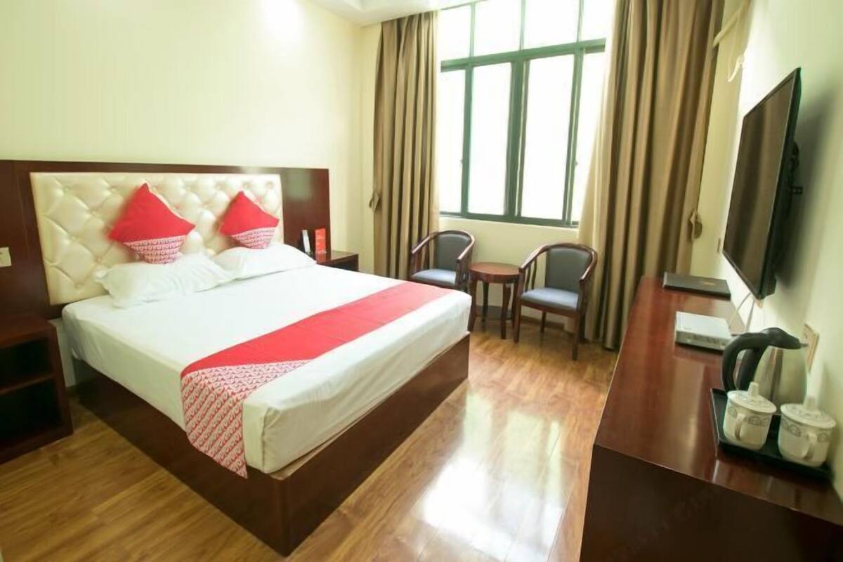 Quarto de hotel com uma cama grande, uma escrivaninha, uma cadeira, uma televisão e uma janela com cortinas.