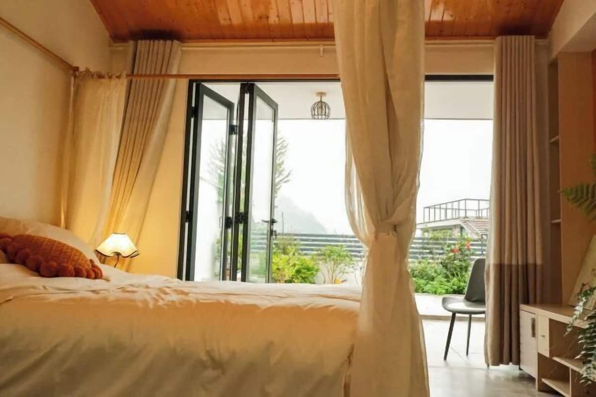 A bedroom with a bed, curtains, a nightstand with a lamp, and a view of greenery outside.