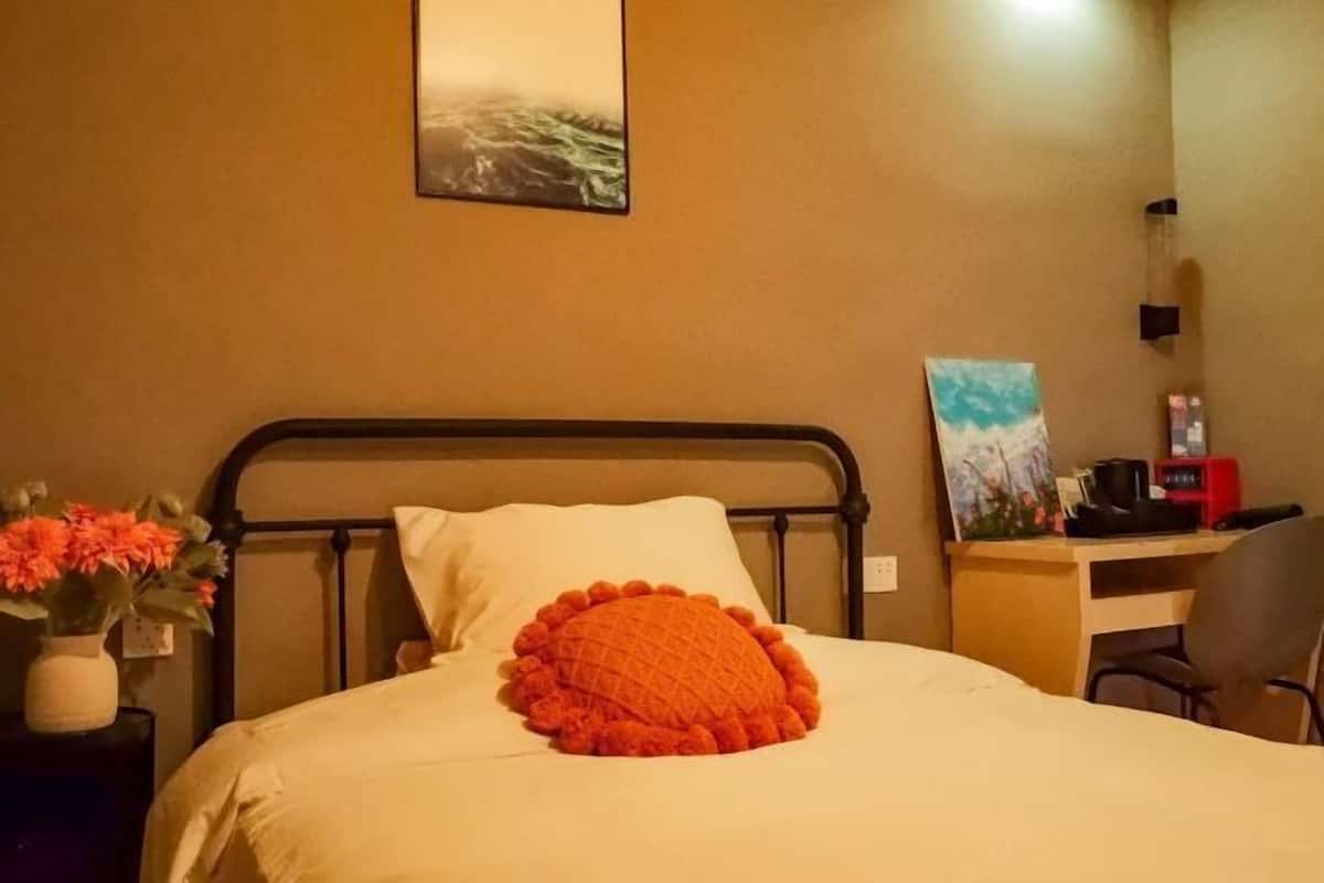 A hotel room with a bed, a desk, a chair, a painting on the wall, and a vase with flowers on the nightstand.