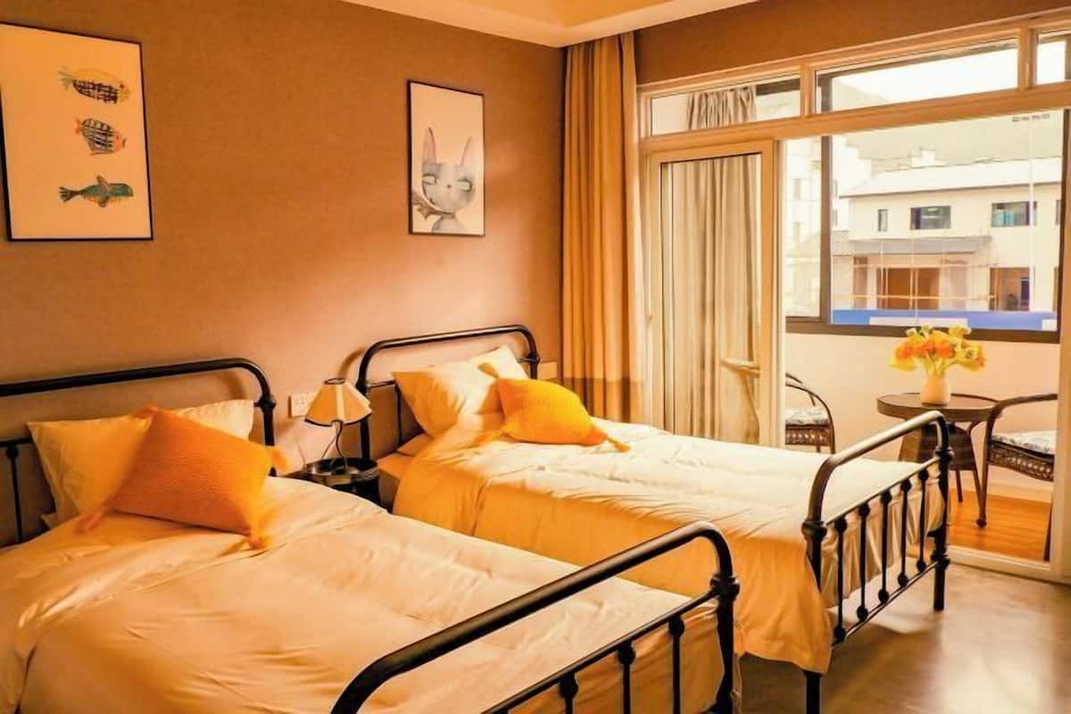 A hotel room with two beds, a window with curtains, a small table with a vase, and artwork on the wall.