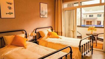 A hotel room with two beds, a window with curtains, a small table with a vase, and artwork on the wall.
