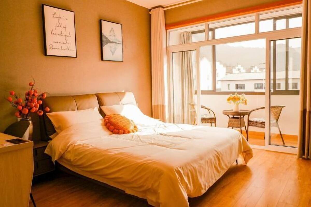 A hotel room with a large bed, a small table with chairs, a vase with flowers, and a balcony with a view of buildings.