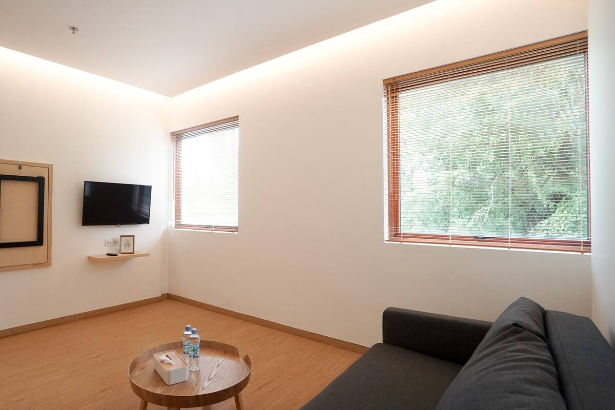 A modern living room with a flat-screen TV, a sofa, a coffee table with a tissue box and water bottles, and two windows with blinds.