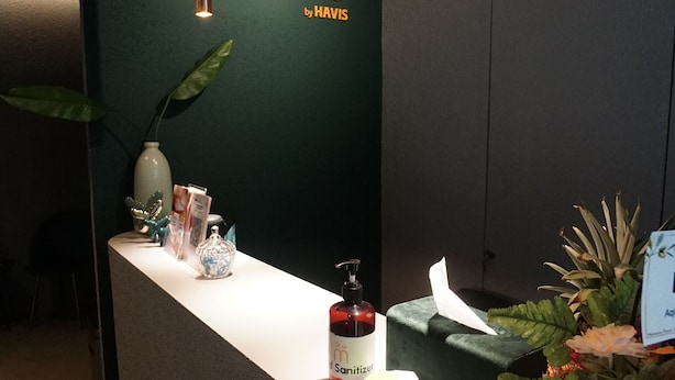 A reception desk with a green wall, a sign reading "manuka by HAVIS", a bottle of sanitizer, a plant, and a vase. - Manuka By Havis Hotel at Penuin (Batam)